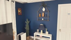 A room with a blue wall, a white door, a white console table with a lamp, a cactus, and a shelf with decorative items. - Zen Cove- Home in West Saint John. 7 minutes from the Irving Nature Park. (Saint John)