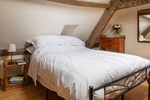 A single bed with white bedding in a room with wooden beams, a nightstand with books, a dresser, and a mirror on the wall. - Tranquility House (Painswick)