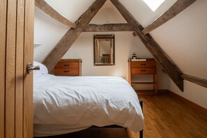 A bedroom with a sloped ceiling, wooden beams, a bed with white bedding, two wooden nightstands, and a mirror. - Tranquility House (Painswick)