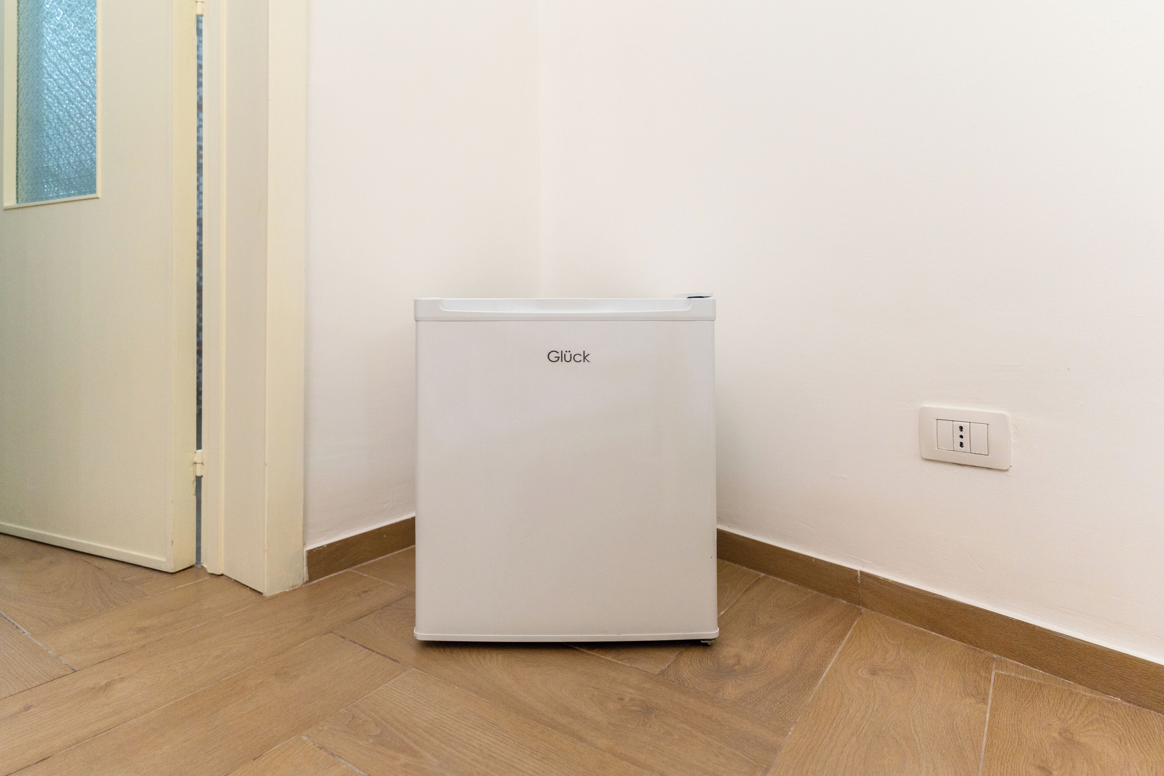 A white Glück refrigerator in a corner of a room with a wooden floor and a white wall.