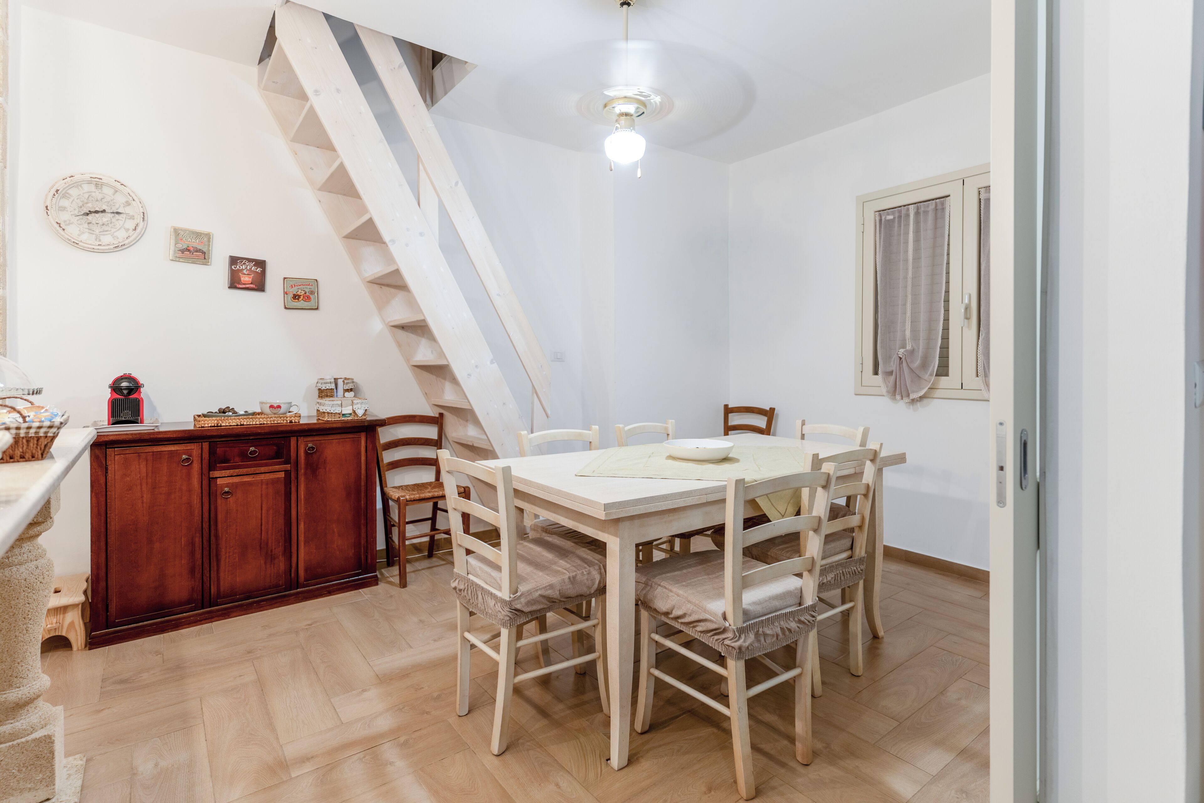A dining area with a wooden table and chairs, a staircase, and a kitchen area with cabinets and a coffee machine.