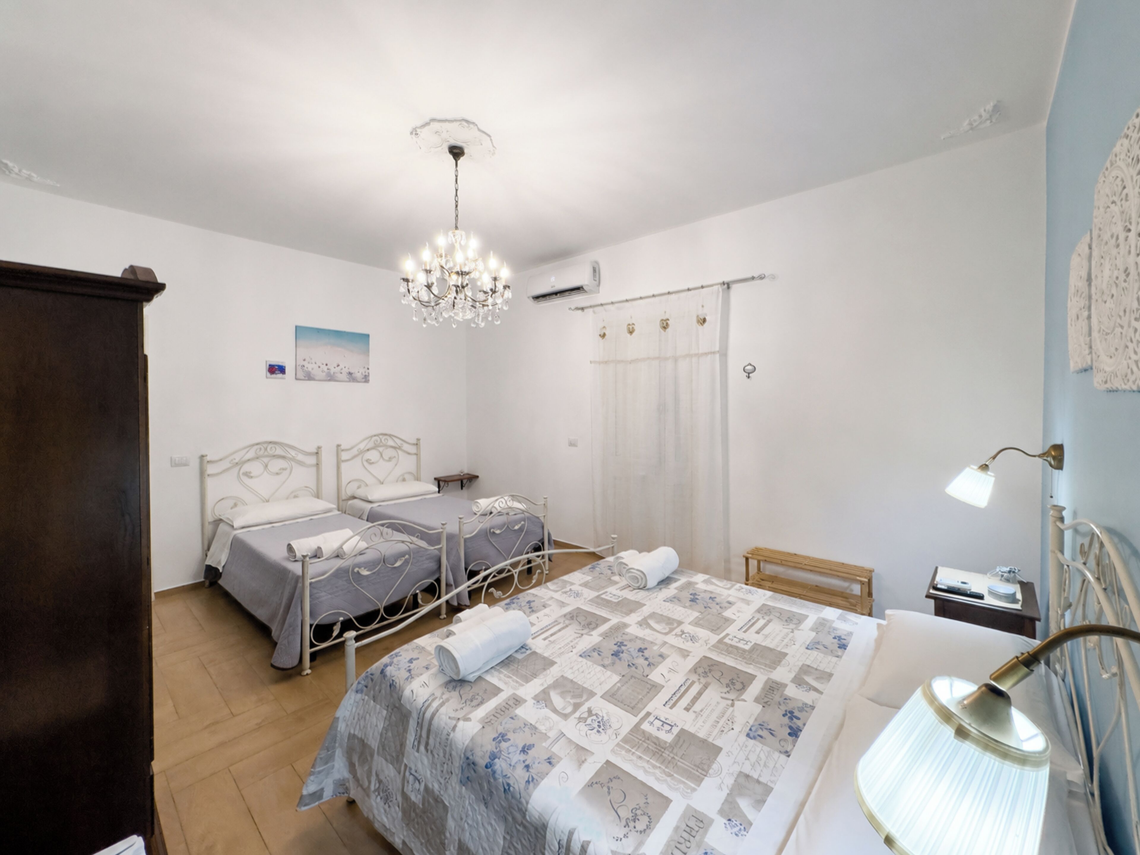 A bedroom with two beds, a chandelier, a bedside table, a lamp, and a window with curtains.