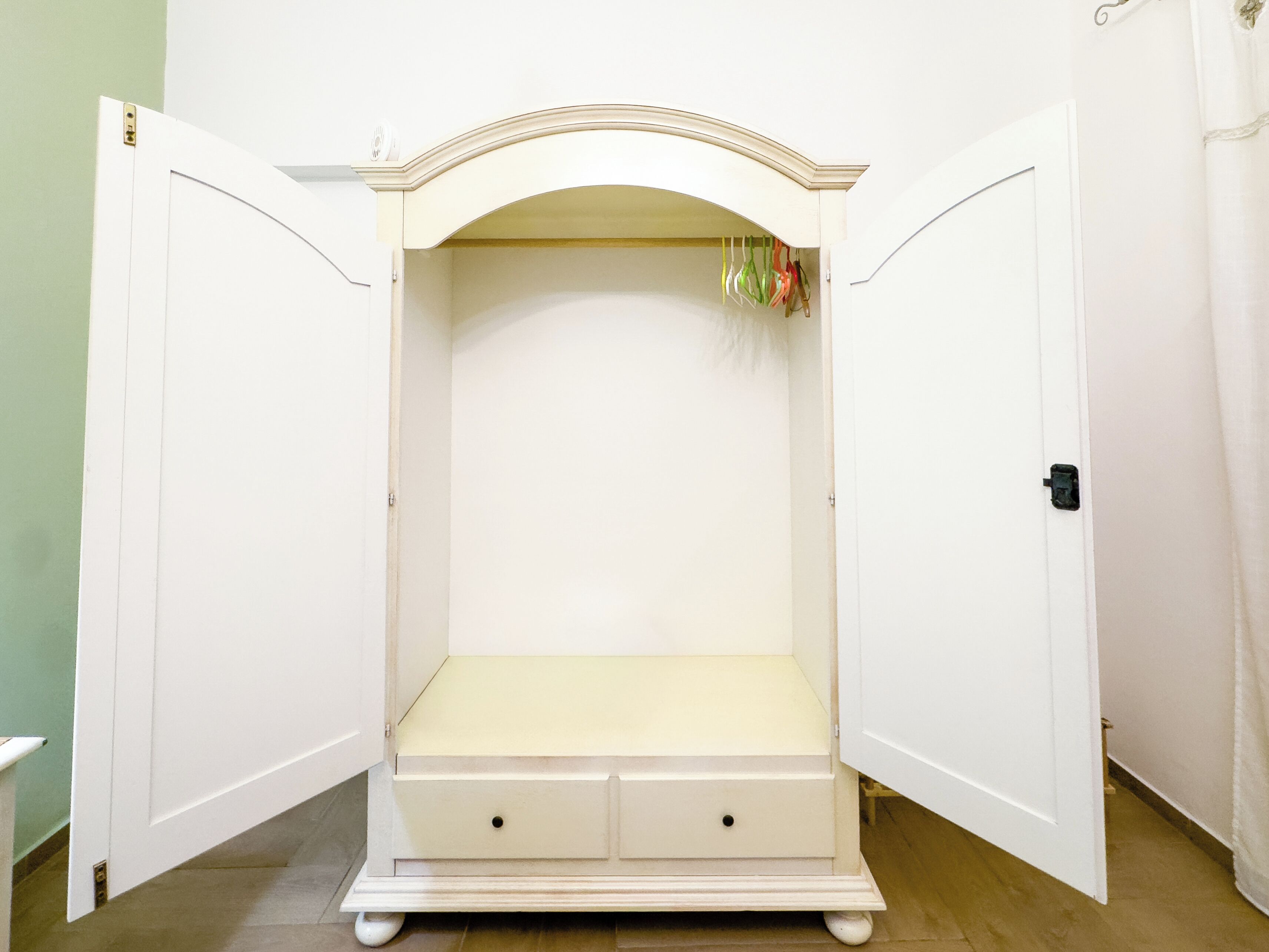 A white armoire with a curved top and two drawers, positioned between two open doors.