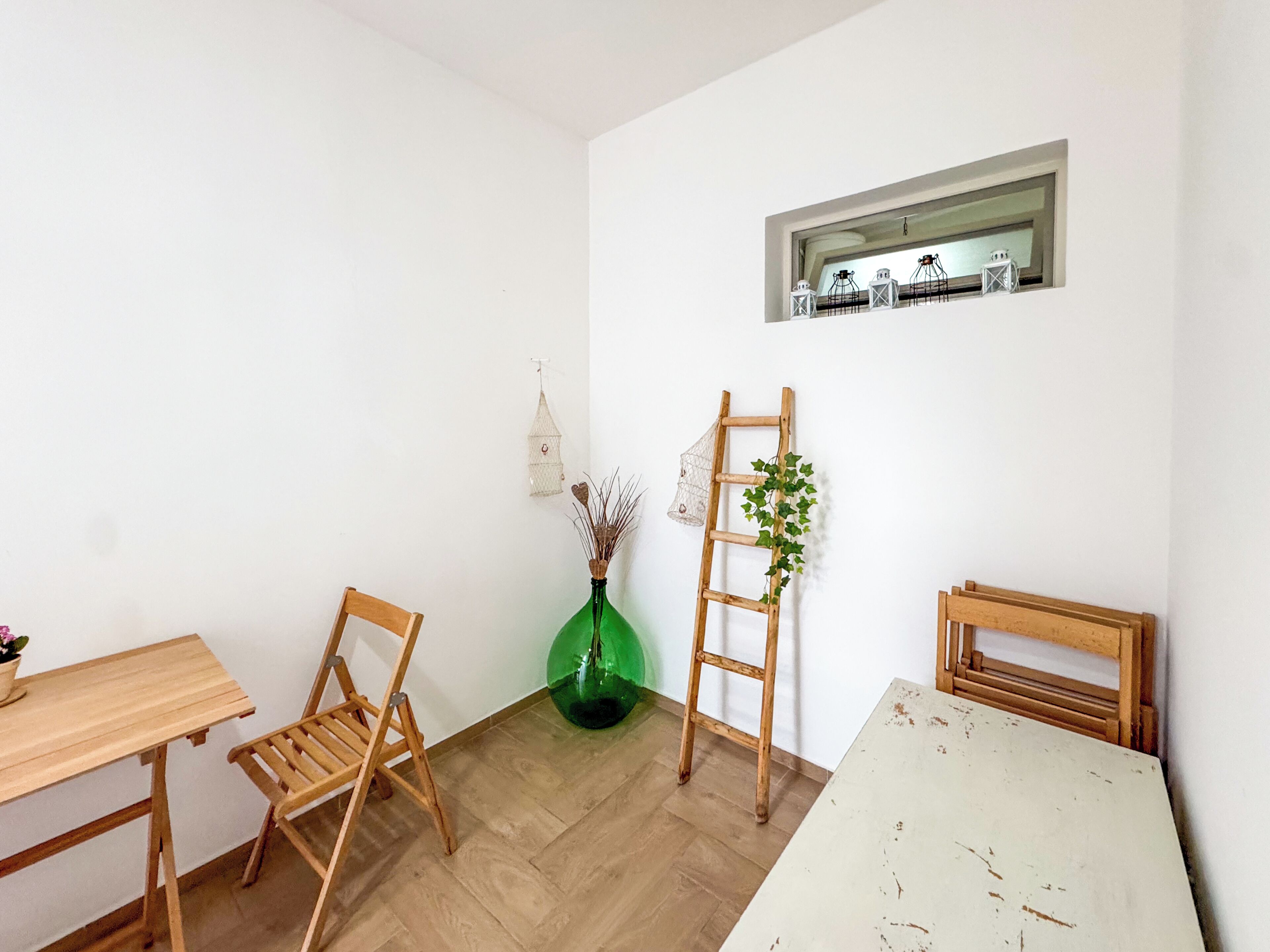A room with a wooden table, two chairs, a ladder, a green vase with plants, and a small shelf with decorative items.