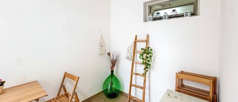 A room with a wooden table, two chairs, a ladder, a green vase with plants, and a small shelf with decorative items.
