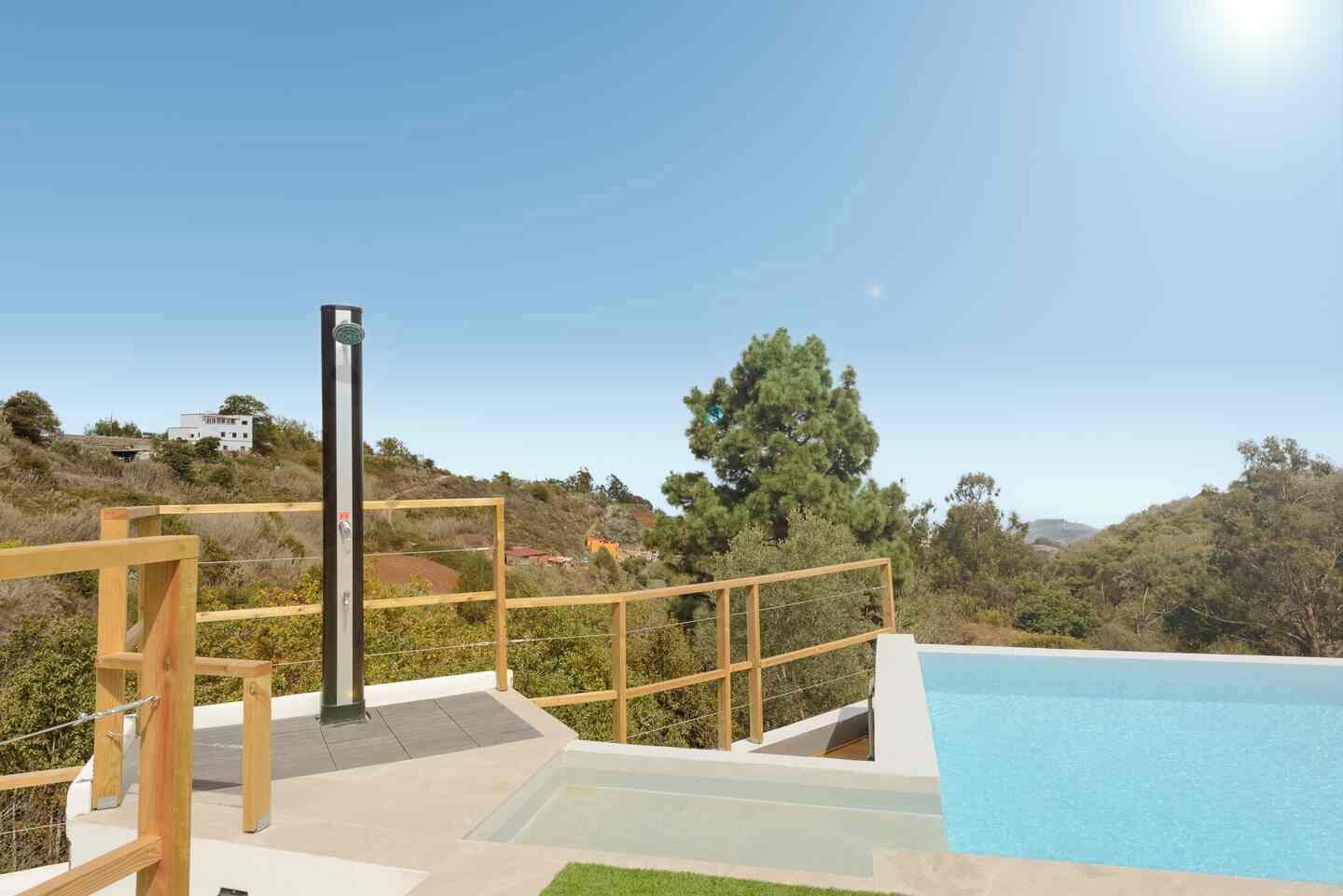 Una piscina con cubierta de madera y un poste de metal, rodeada de follaje y colinas.