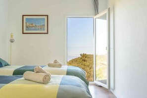 A bedroom with two beds, a framed painting, and a large window with a view of a hill.