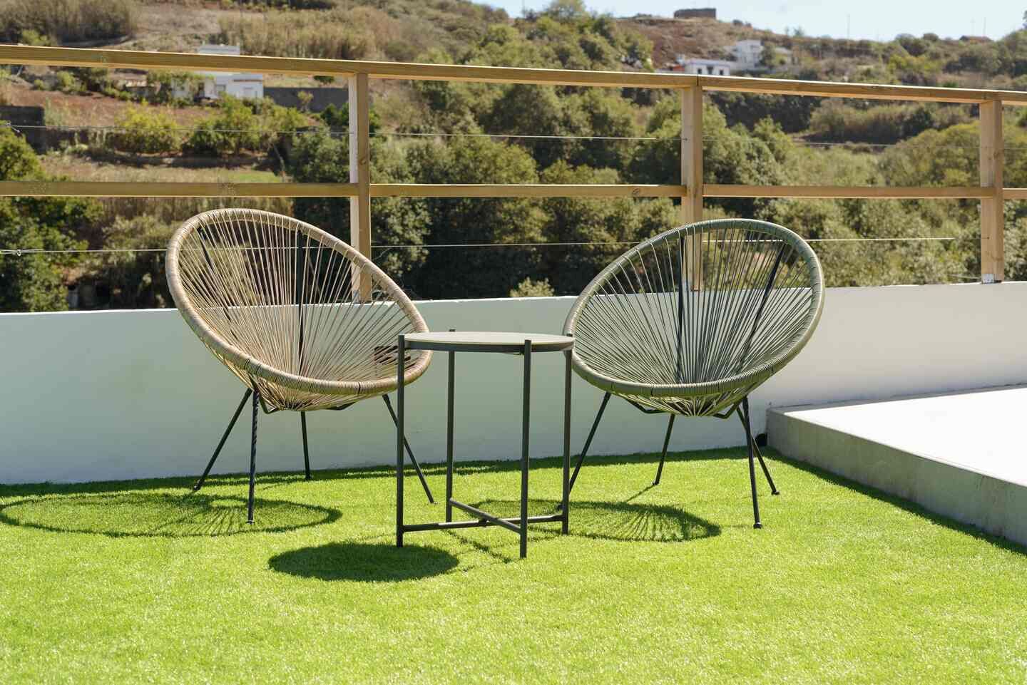 Duas cadeiras de vime com estrutura de metal em um terraço com grama sintética, com vista para uma paisagem acidentada.