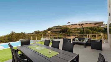 An outdoor patio with a dining table, chairs, and a sofa. There is a pool in the background, and the area overlooks a hilly landscape.