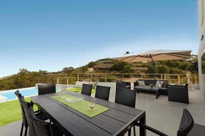 An outdoor patio with a dining table, chairs, and a sofa. There is a pool in the background, and the area overlooks a hilly landscape.