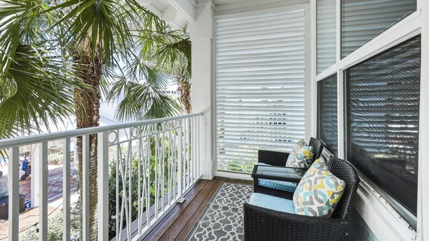 En balkon med gelænder i hvidt, en fletsofa med mønstrede puder og udsigt til palmer og bygninger.
