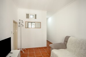 Une pièce avec un mur blanc, une porte, une petite fenêtre, un miroir, un canapé gris et un téléviseur à écran plat.