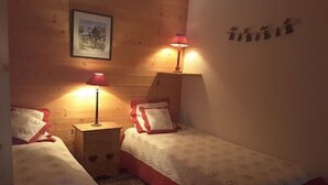 A room with two beds, each with a red pillow and a patterned bedspread. There are two bedside tables with lamps, and a framed picture on the wall. - Meribel Mottaret Apartment at the Foot of the Slopes, Sleeps 5, All-Inclusive (Les Allues)