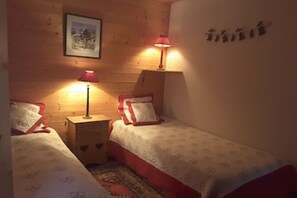 Ein Zimmer mit zwei Betten, jedes mit einem roten Kissen und einer gemusterten Tagesdecke. Es gibt zwei Nachttische mit Lampen und ein gerahmtes Bild an der Wand.