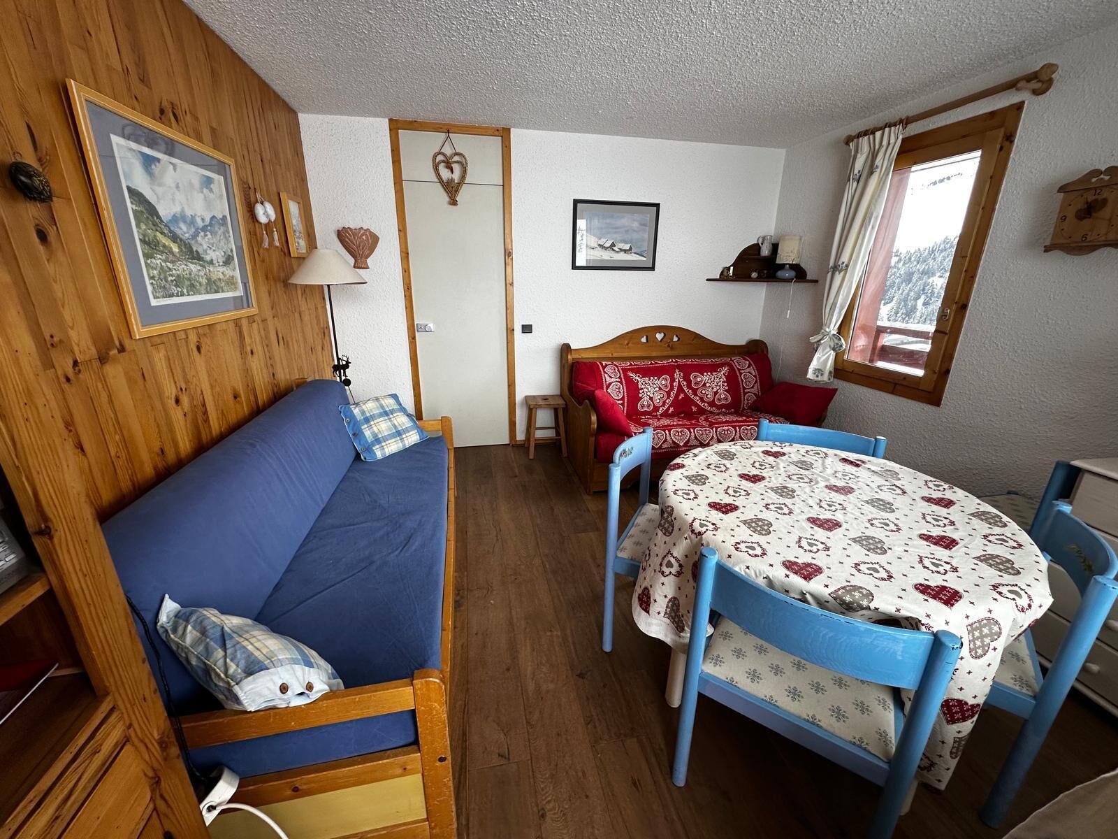 A cozy room with a blue sofa, a wooden bed, a small table with a patterned tablecloth, and a window with curtains.