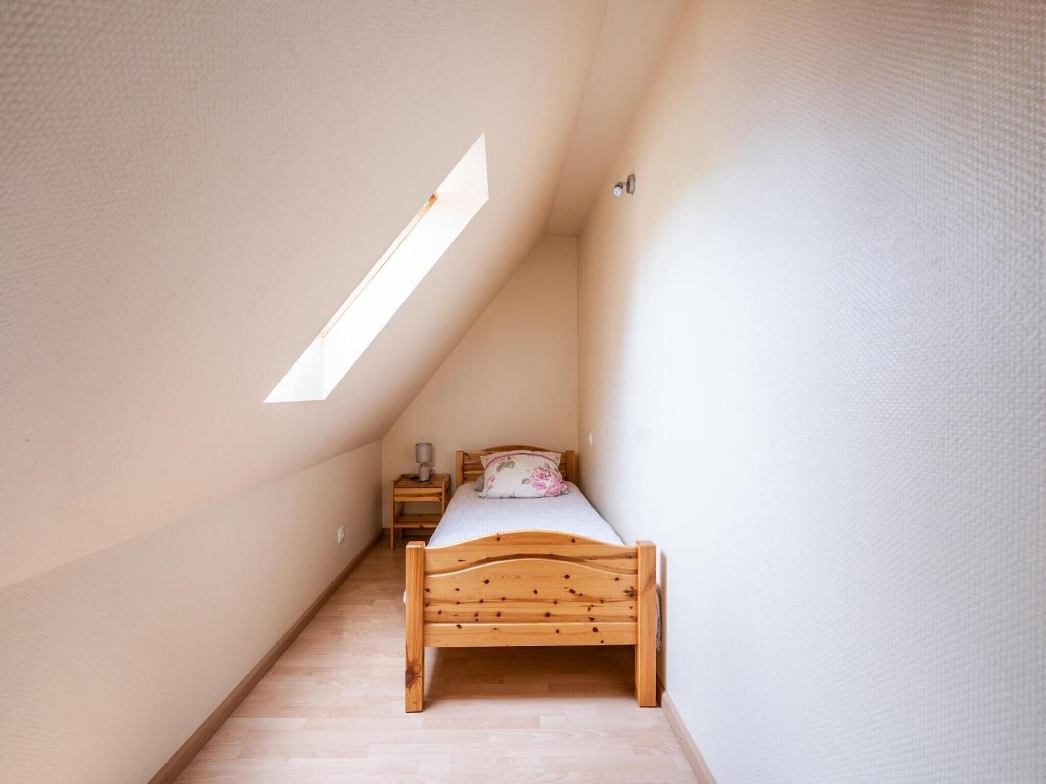 A small, narrow room with a sloped ceiling, a wooden bed, a nightstand, and a wall-mounted light fixture.