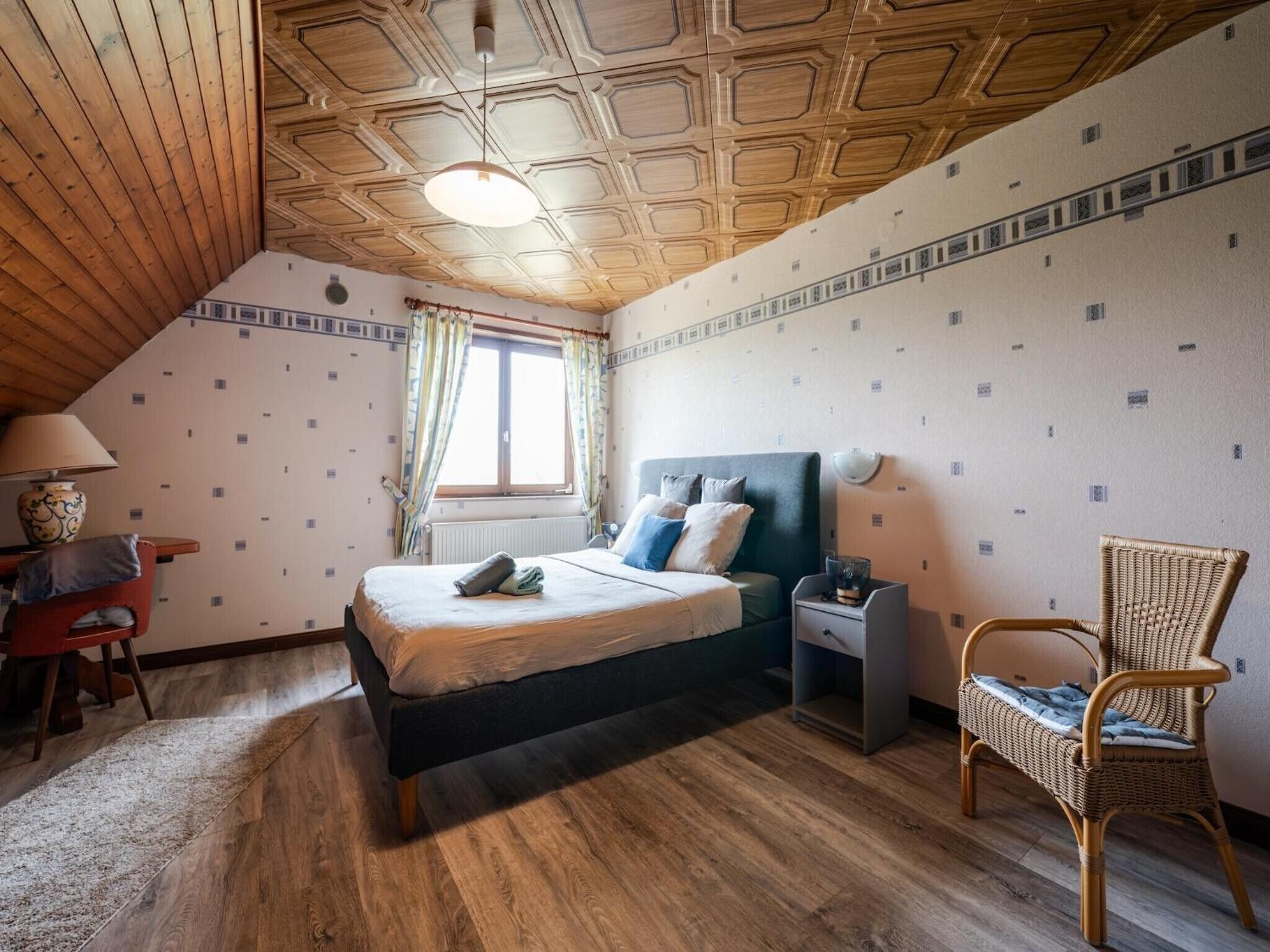 Une chambre à coucher avec un plafond en bois, un lit avec des oreillers, une chaise, un bureau et une fenêtre avec des rideaux.