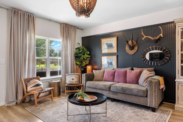 A living room with a sofa, chairs, a coffee table, and a large window with curtains. - Apple Street Getaway - Fire Pit & Close to Main St (Fredericksburg)