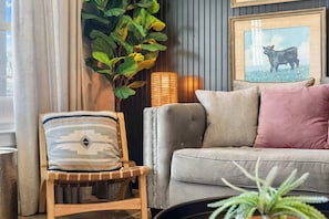 A living room with a grey sofa, a striped pillow, a wooden chair, a large plant, a lamp, and a framed picture on the wall. - Apple Street Getaway - Fire Pit & Close to Main St (Fredericksburg)