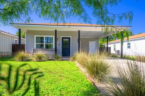 A single-story house with a front porch, a door, and a small front yard with grass and shrubs. - Apple Street Getaway - Fire Pit & Close to Main St (Fredericksburg)