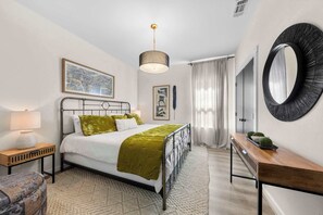 A bedroom with a bed, bedside tables, a mirror, and artwork on the walls. - Apple Street Getaway - Fire Pit & Close to Main St (Fredericksburg)