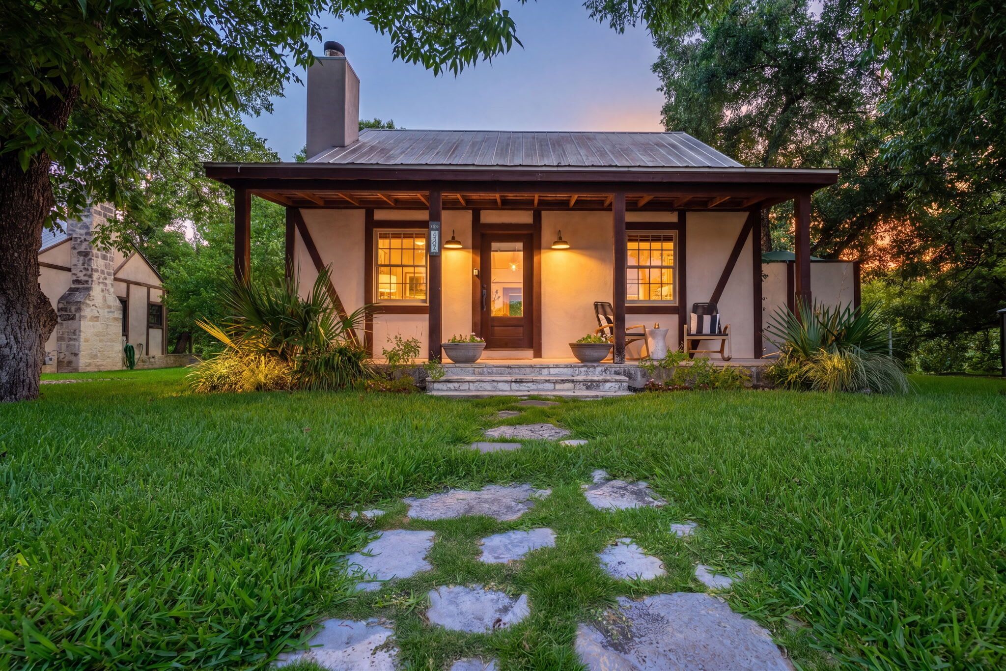 A two-story house with a stone chimney, a front porch with two chairs, and a stone pathway leading to the entrance.