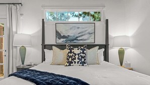 Una cama bien hecha con un edredón azul, dos lámparas de noche, una ventana con vistas a árboles y un cuadro en la pared.