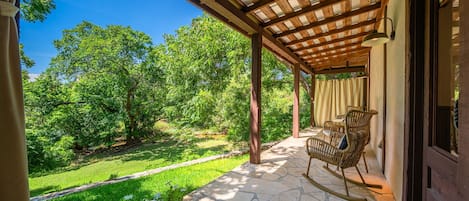 A covered patio with stone flooring, a rocking chair, and a view of a lush green landscape.