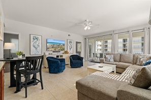 Ein großzügiges Wohnzimmer mit einer Couch, Sesseln, einem Couchtisch und einem Fernseher. Ein Balkon bietet Blick auf die Stadt.