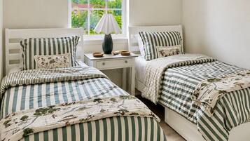 Quarto com duas camas de solteiro, uma mesinha de cabeceira com abajur, uma janela com persianas e um objeto decorativo na parede.