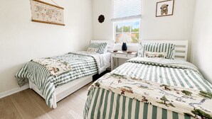 Un dormitorio con dos camas individuales, una ventana con persianas, una mesita de noche con lámpara y un adorno en la pared.