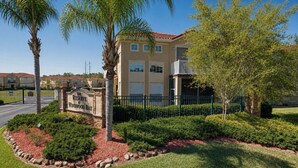 Edificio residencial con entrada protegida por un portón, rodeado de jardines ajardinados y palmeras.