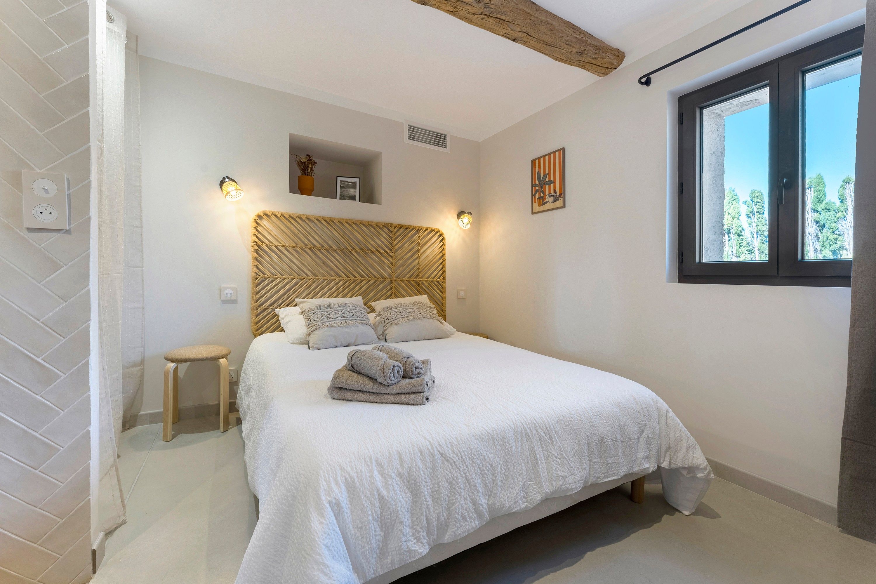 Un dormitorio con cama, mesita de noche, lámpara de pared y ventana.