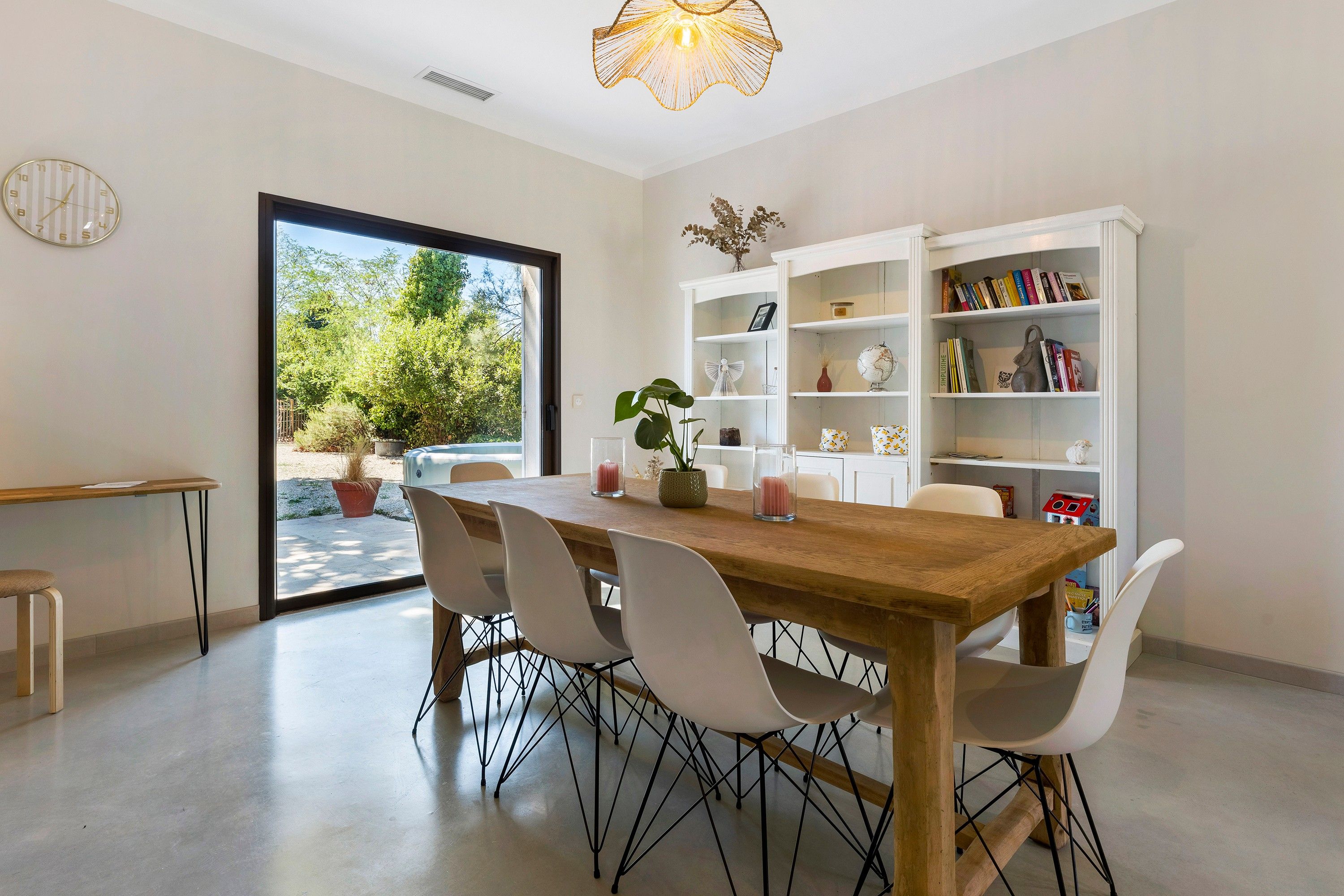 Un coin repas avec une table en bois, des chaises blanches, une bibliothèque et une vue sur la verdure à l’extérieur.
