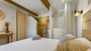 Quarto com cama grande, vigas de madeira, parede de tijolos, porta de madeira e luminária fixada na parede.