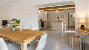Uma zona de refeições moderna, com uma mesa de madeira e cadeiras brancas, cozinha ao fundo e um ambiente acolhedor.