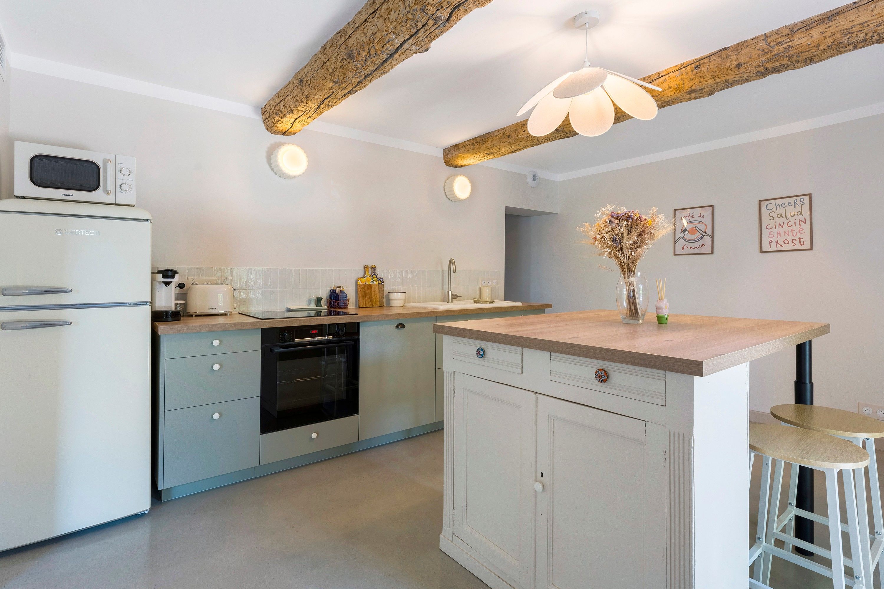 A modern kitchen with a built-in oven, a microwave, and a sink. There is a wooden ceiling beam and pendant lighting.
