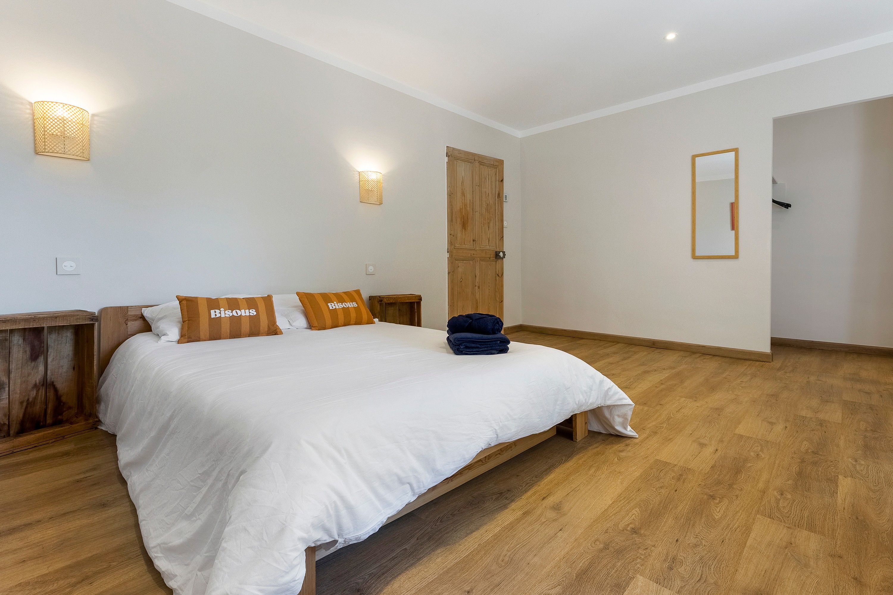 Une chambre avec un grand lit, une tête de lit en bois, deux lampes fixées au mur, une porte en bois et un miroir sur le mur.