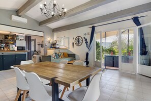 Uma área de refeições com mesa de madeira e cadeiras brancas, cozinha ao fundo e porta de vidro deslizante para área externa.