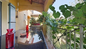 A covered outdoor balcony with a glass table, red vase, and potted plants.