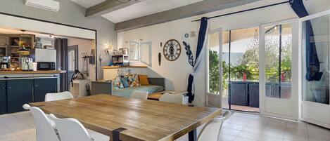 A dining area with a wooden table and white chairs, a kitchen in the background, and a sliding glass door leading to an outdoor area.