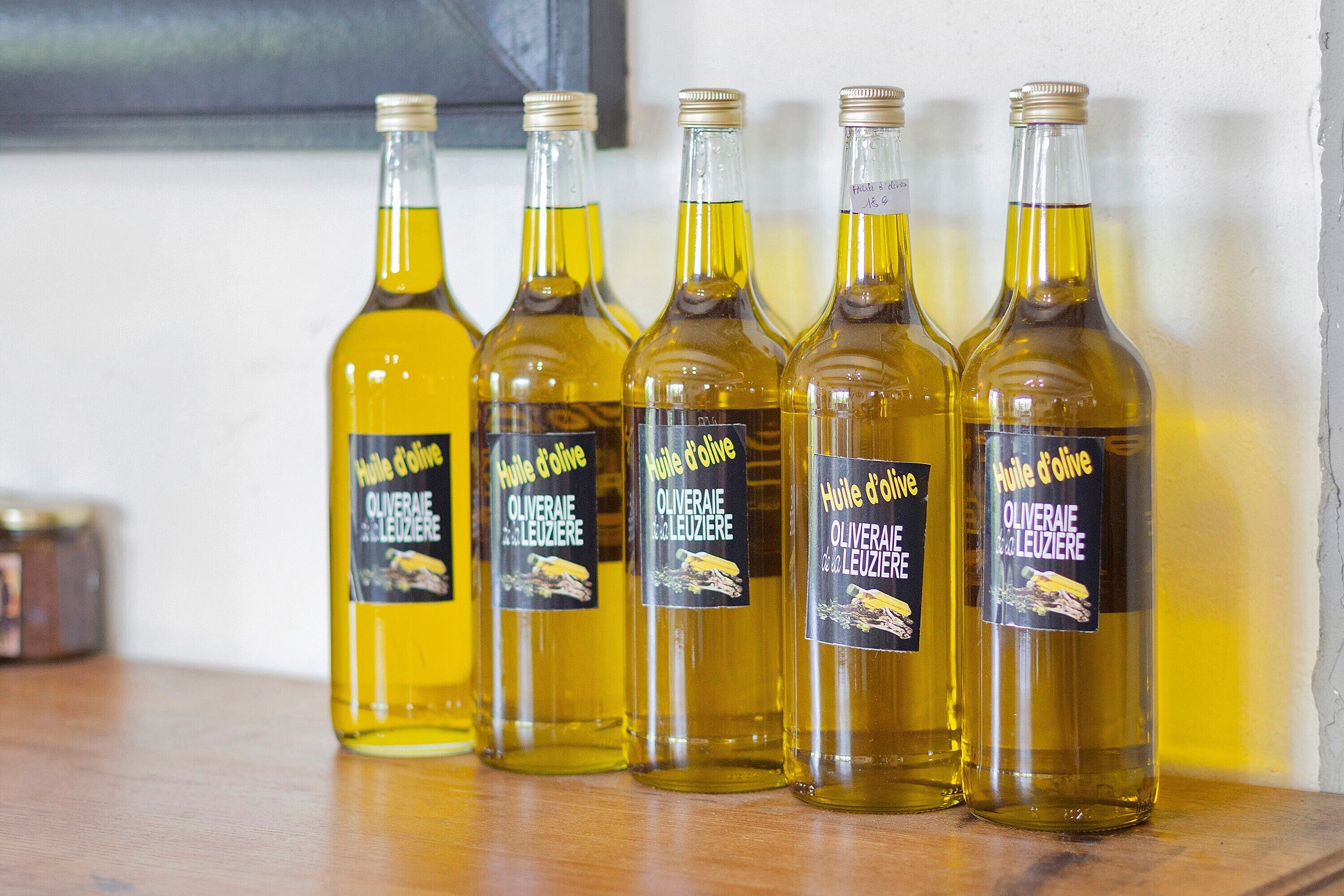 Cinq bouteilles d’huile d’olive, toutes étiquetées, sont posées sur une surface en bois devant un mur uni.