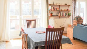 Uma área de refeições com mesa, cadeiras e vista para o exterior. Há prateleiras com livros e um pequeno modelo de construção.