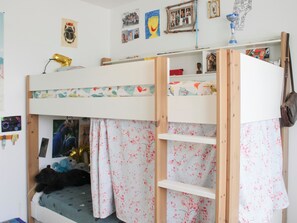 Ein Etagenbett, wobei die untere Koje mit einem blumigen Vorhang versehen ist, ein Regal mit diversen Gegenständen und eine Wand, die mit Fotos und einer Karte dekoriert ist.
