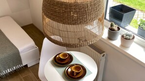 Een moderne kamer met een witte tafel, twee kopjes, een geweven lamp en een raam met plantenpotten.