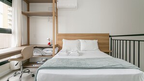 Una habitación de hotel con cama, escritorio, silla, estante con toallas y ventana con cortinas.