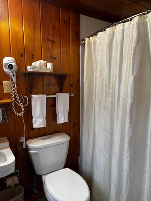 Un baño con pared de madera, un inodoro blanco, un secador de pelo en un estante y una cortina de ducha.