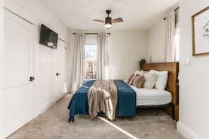 Un dormitorio con cama, ventilador de techo, ventana con cortinas y televisor montado en la pared.