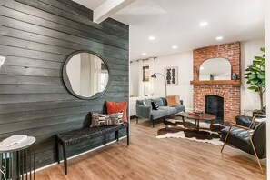 A modern living room with a dark wooden wall, a round mirror, a sofa, a coffee table, a leather armchair, a brick fireplace, and a cowhide rug. - Spacious & Historic 3BR Near German Village. Fire Pit, Yard, Grill.Veteran-Owned (Columbus)