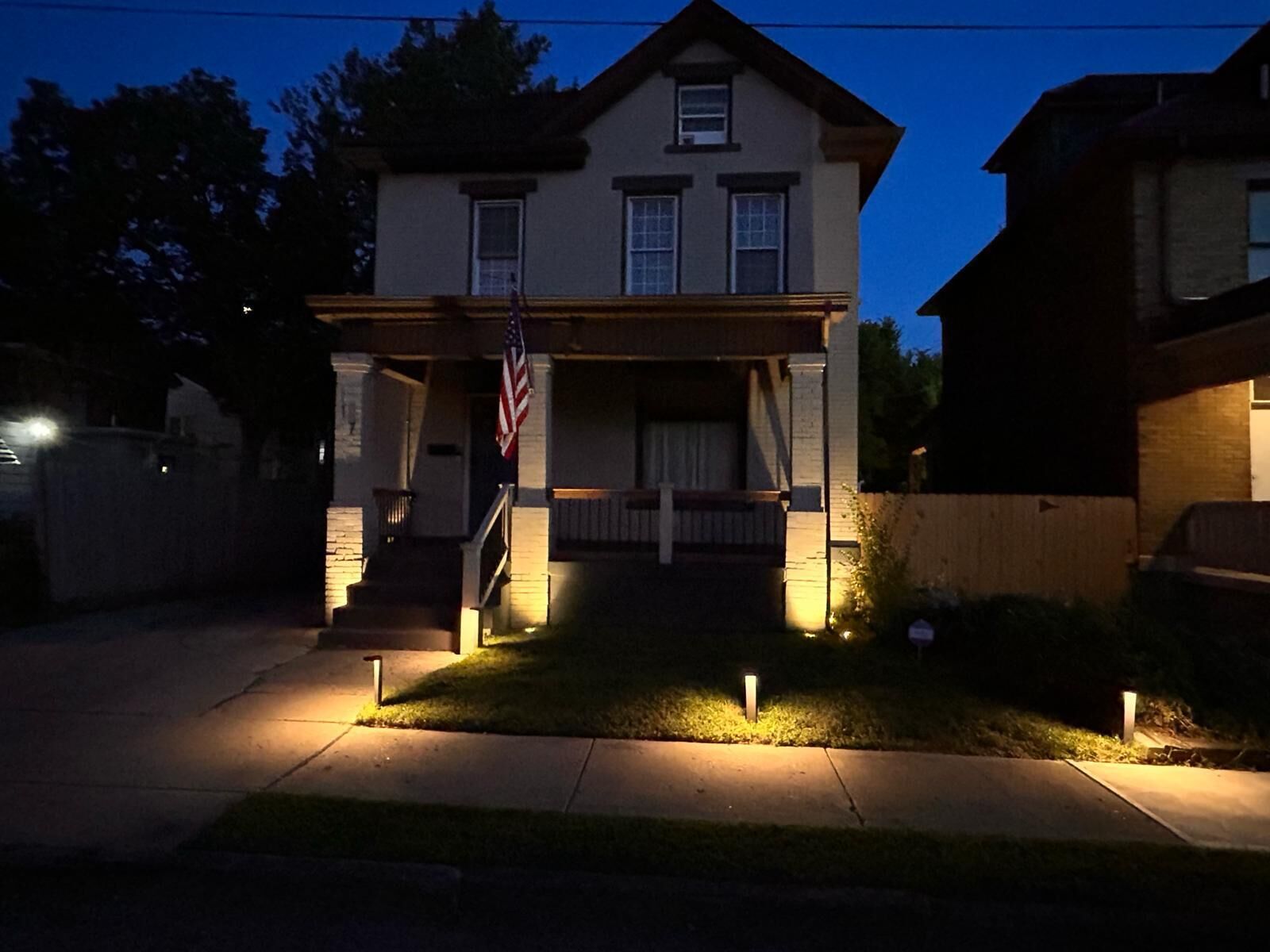 Una casa de dues plantes amb un porxo, una bandera estatunidenca i un jardí ben il·luminat a la penumbra.